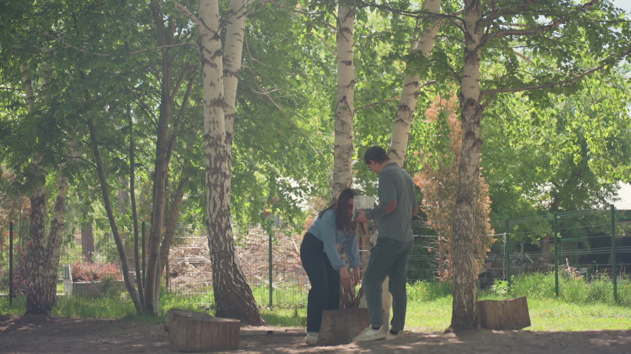 두 사람이 야외의 자연 통나무 위에서 흰색 개를 훈련시키고, 햇빛 아래 숲 속 통나무 위에서 개 민첩성 훈련을 합니다. 두 사람은 햇빛과 나무가 있는 곳에서 흰색 개가 통나무 위에서 균형을 잡도록 가르칩니다.