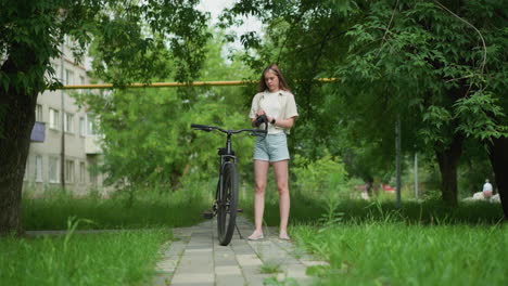 woman holds black gloves as she approaches parked bicycle on paved path, preparing to wear gloves while smartwatch is visible on right hand, background includes and people passing by in distance