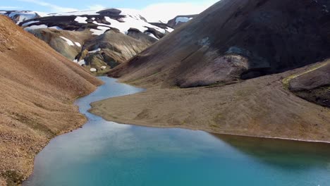 flight over scree and a blue lake in the valley gradually revealing rainbow mountains and white river in the gorge between orange steep slopes