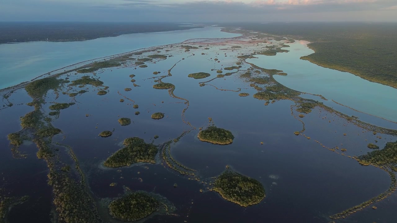 Imagem de drone voando para a frente sobre a vasta Lagoa de Bacalar, revelando ilhotas de mangue abstratas e padrões de zonas úmidas espalhados sobre águas calmas e reflexivas no México