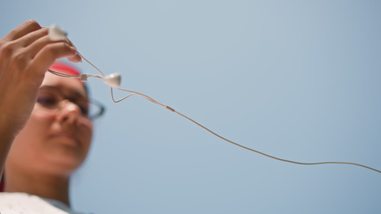 Young woman untangles earphones outdoors, Female adjusting earbuds beneath blue sky cheerful expression, Young woman in red cap happily untangles her earbuds under sunny summer sky in street