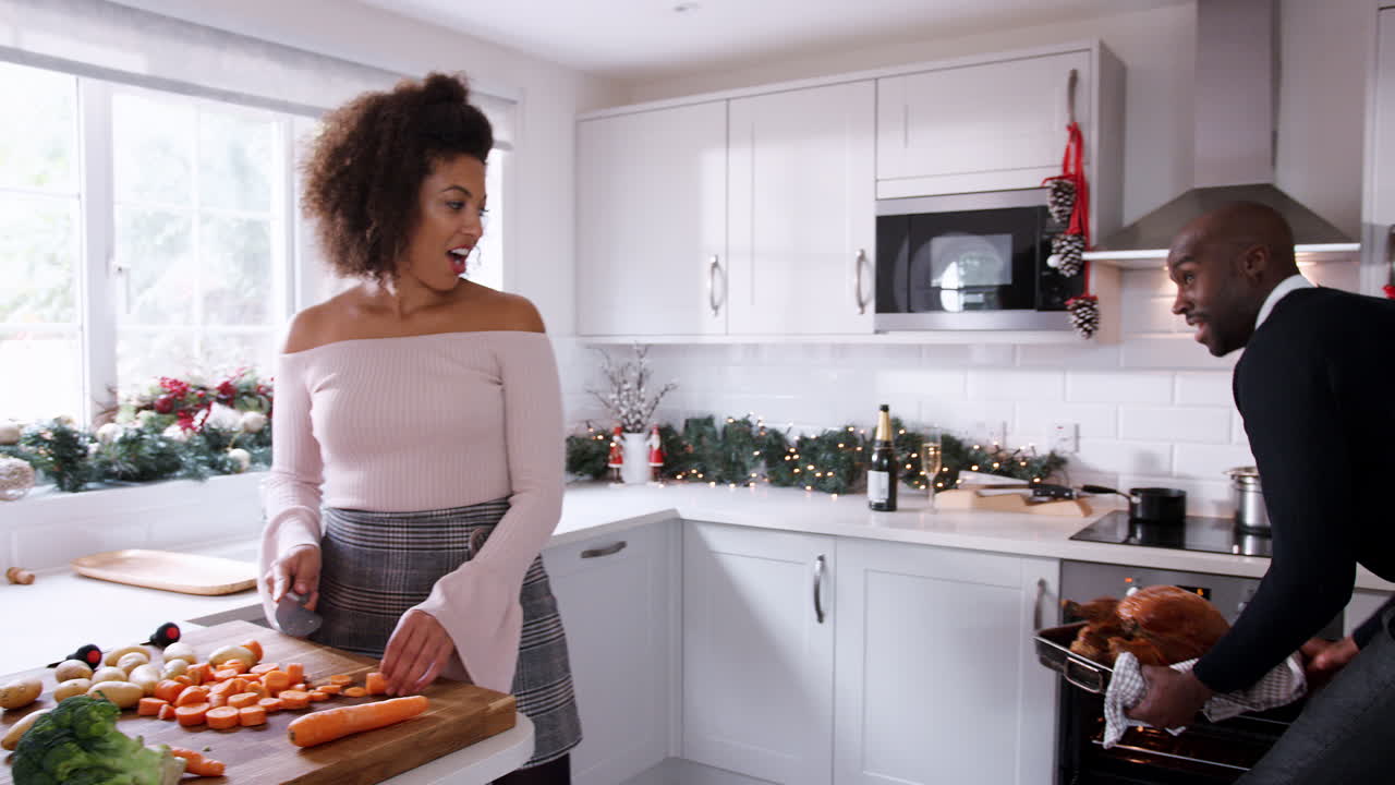 Young adult mixed race couple preparing Christmas dinner together at home, woman cutting vegetables and man basting roast turkey, front view, close up