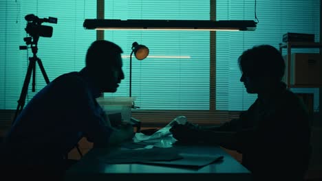Silhouette Video of a Perpetrator or Prisoner Sitting in the Interrogation Room in front of the Silhouette Video of a Perpetrator Prisoner Sitting in the Interrogation Room in front of the Detective the Policemnan Pulls a Knife from Evidence Box Talks to the Suspect True Crime Documentary