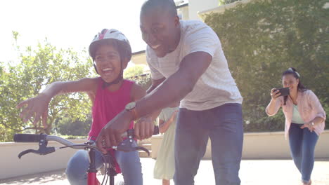 teaching child to ride bike, father supporting while family cheers and takes photos
