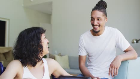 Happy-biracial-woman-in-wheelchair-and-male-partner-looking-at-each-other-and-smiling-in-living-room