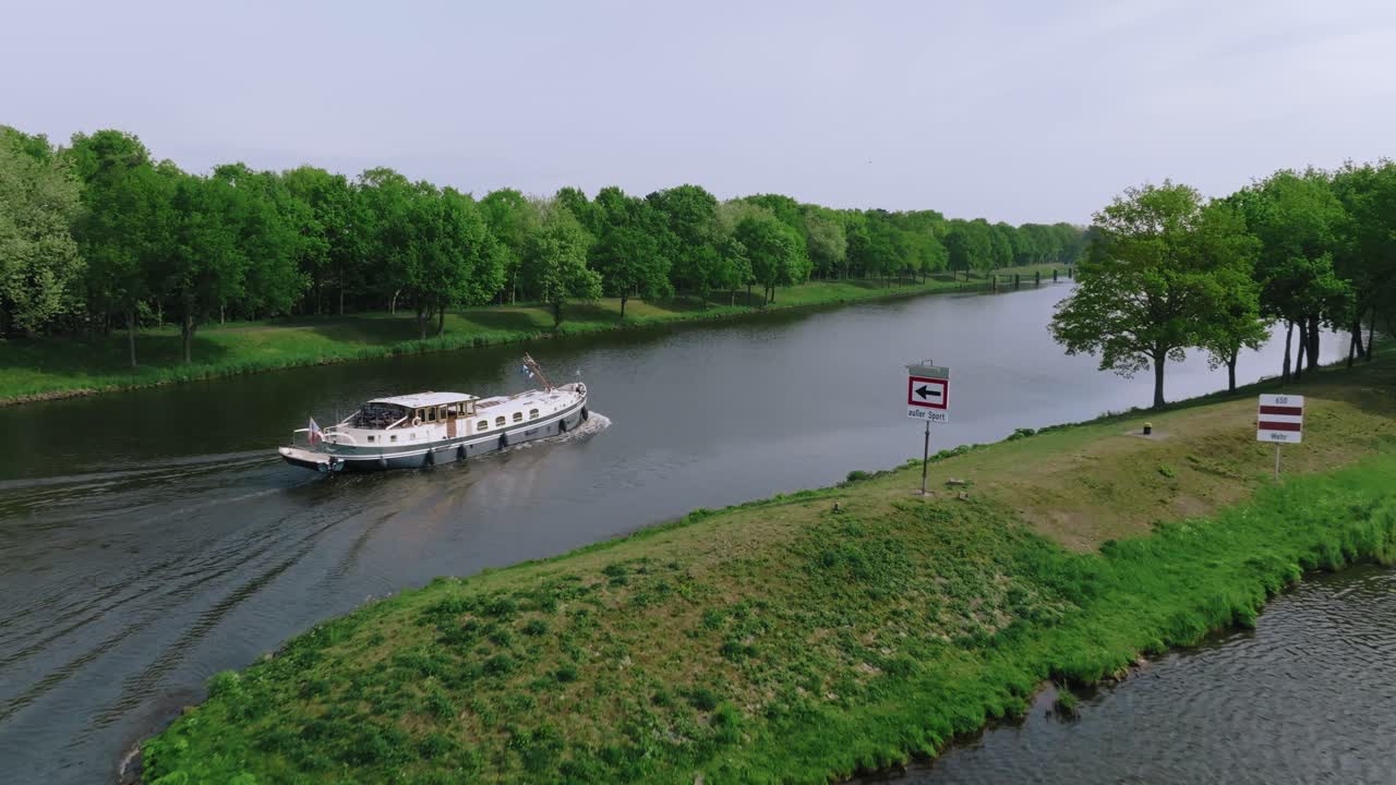 Zoom in aerial shot of a white boat passing a green island on a calm river under soft daylight.