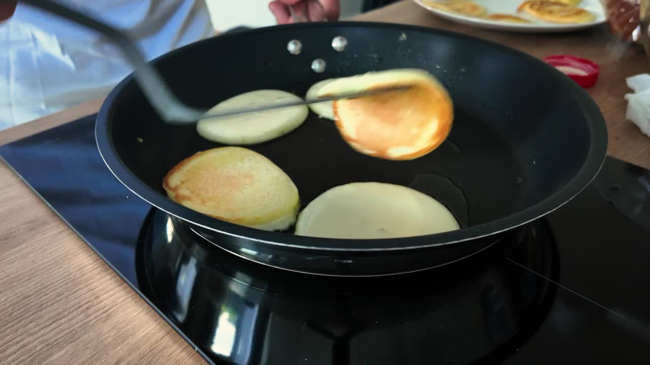 Close-up of a person flipping fluffy pancakes in a non-stick frying pan on an electric stovetop, capturing the process of cooking breakfast in a home kitchen setting