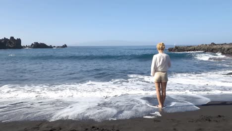 lonely woman standing on beach while ocean waves breaking on black sand coast of tenerife, canary island, spain