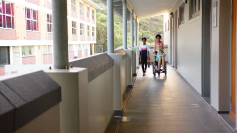 pushing wheelchair, two girl students helping friend in school hallway, smiling together