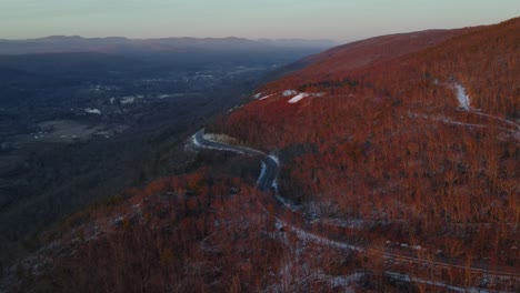 Flying-over-a-scenic-highway-on-a-mountain-high-above-a-vast-valley-during-winter-just-after-sunset,-with-a-little-snow-on-the-ground