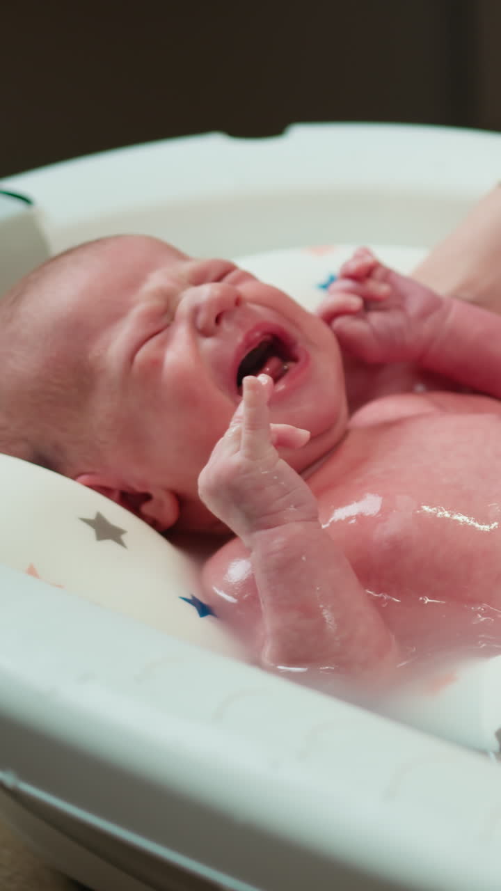 Un bebé recién nacido angustiado llora fuerte mientras lo bañan en una pequeña bañera con unas manos que lo sujetan suavemente