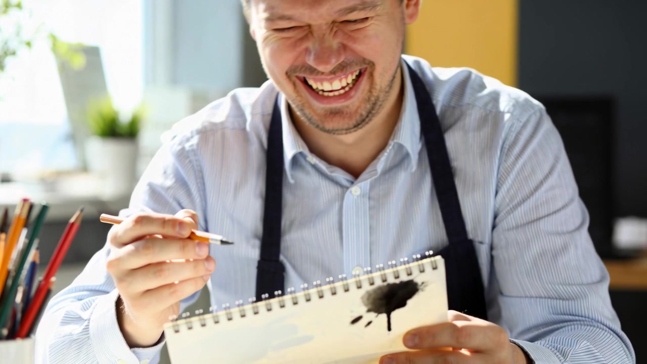 Notando una macchia d'inchiostro, un artista picchietta il pennello alla scrivania con un quaderno da disegno, ridendo in camicia e grembiule.
