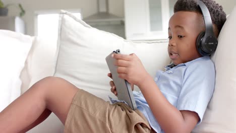 Feliz-Niño-Afroamericano-Con-Auriculares-Tumbado-En-El-Sofá-Usando-Una-Tableta-En-Casa,-Cámara-Lenta