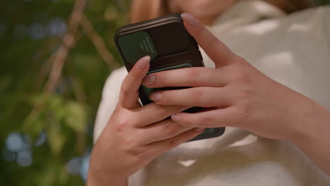 close-up of person operating smartphone with polished nails, typing shadows create a gentle pattern on clothing, background shows natural green surroundings with leaves swaying softly in breeze