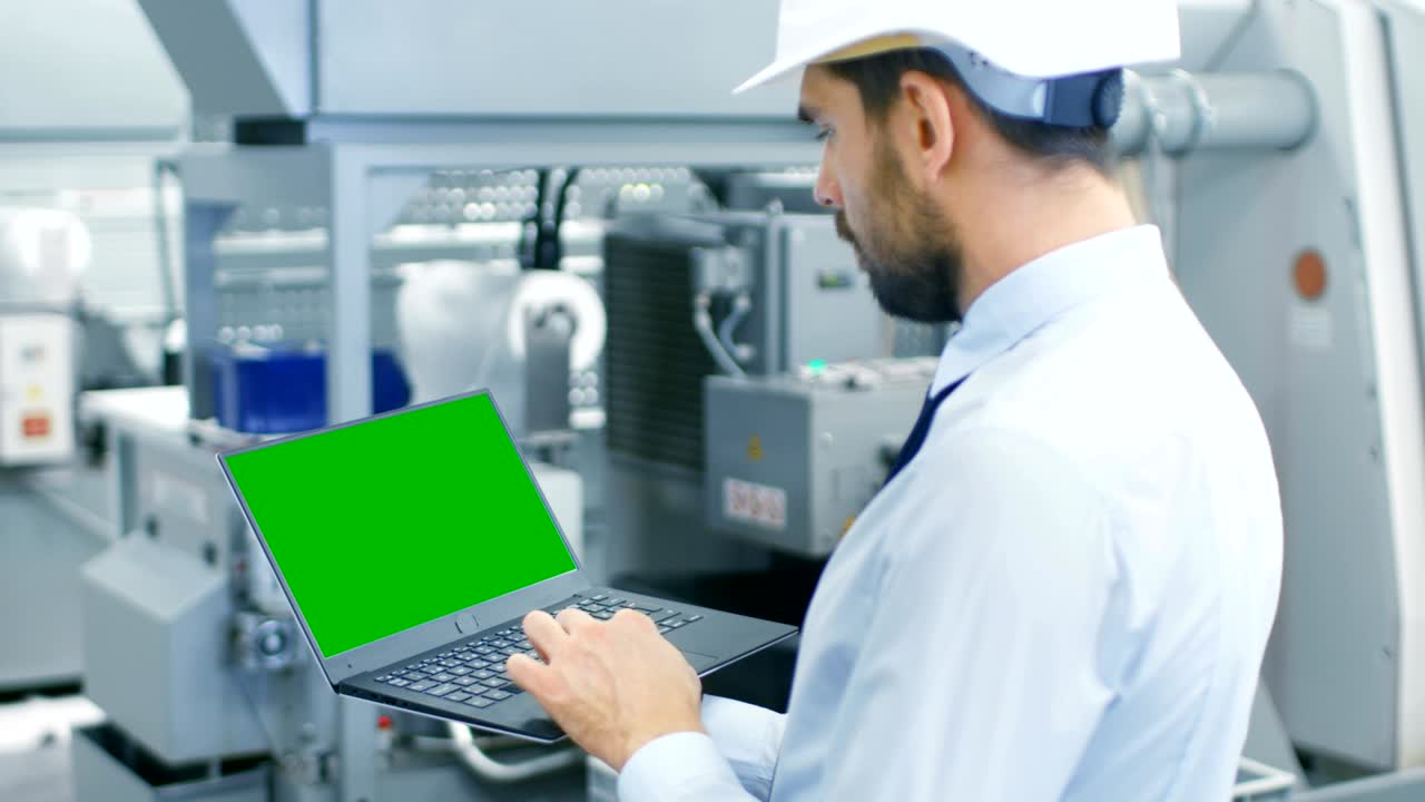 промышленный инженер в жесткой шляпе работает на зеленом модельном экране ноутбука, стоя в середине современной фабрики по производству электроники.