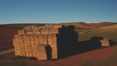 Aerial-shot-flying-towards-and-around-a-big-block-of-stacked-straw-bales-near-a-road-on-green-and-brown-plowed-fields