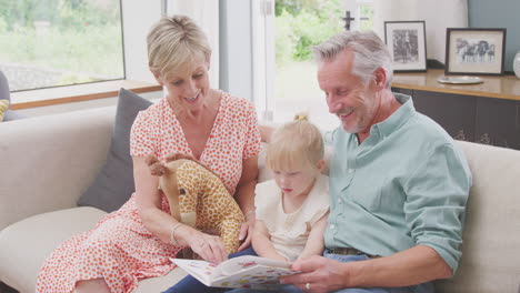 grandparents sitting on sofa with granddaughter at home reading book together