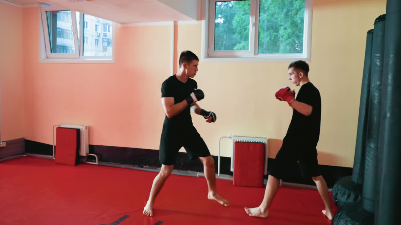 Boxers sparring barefoot on red mat inside training gym, one delivering straight punch while opponent defends with gloves up, showcasing combat practice, discipline, focus, and determination