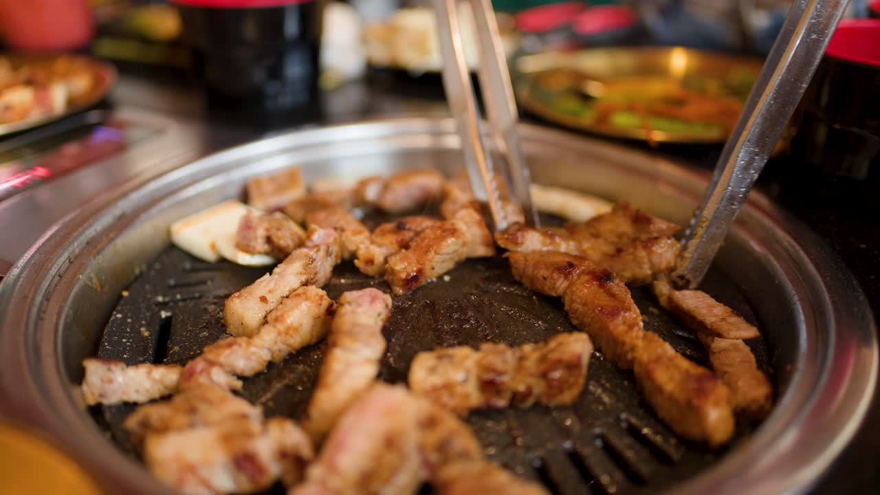 Close-up of pork belly grilling on tabletop pan, tongs turning meat in warm restaurant lighting