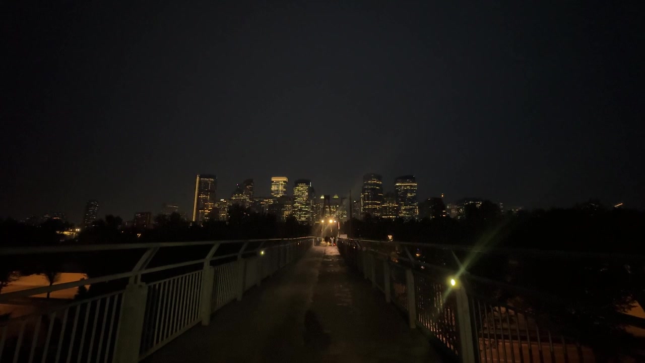 Horizonte de arranha-céus da cidade visto de uma passarela com pedestres, à noite, em formato amplo.
