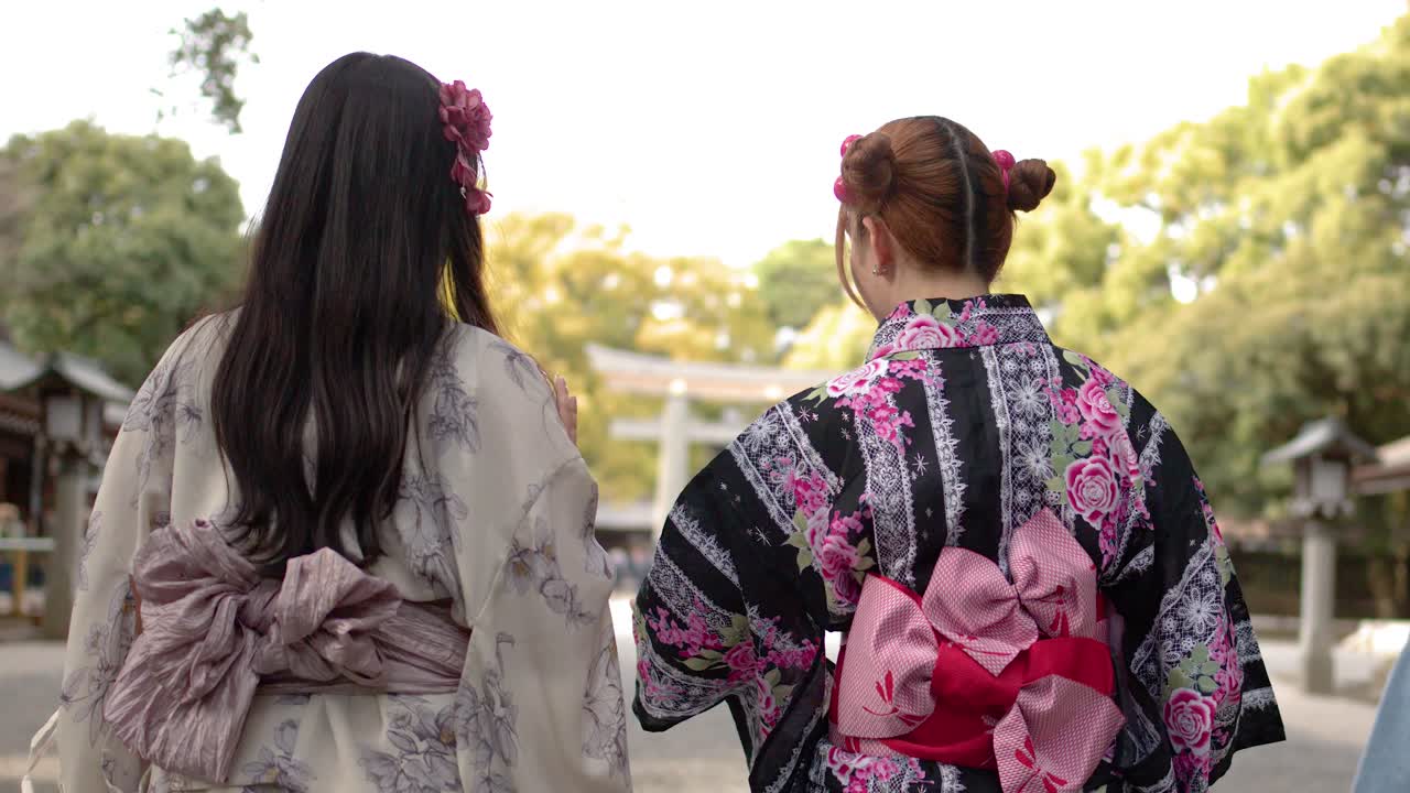 Ảnh chụp từ phía sau hai người phụ nữ mặc kimono họa tiết hoa đi bộ qua một công viên đền thờ Nhật Bản.