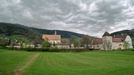 historic monastery with white walls and steep roofs set in green fields, surrounded by alpine hills and cloudy skies, in a tranquil village landscape typical of rural austria shot from moving vehicle
