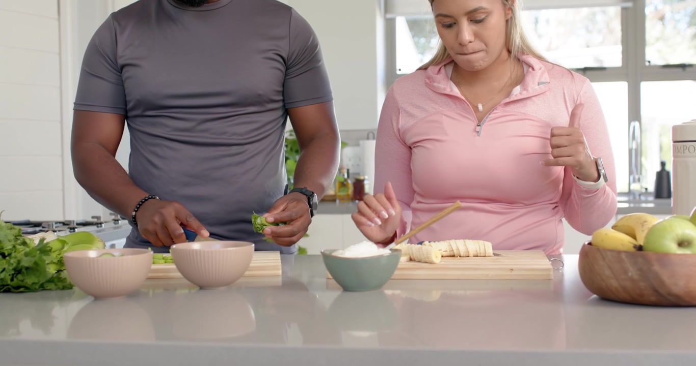 Coppia afroamericana che prende un contenitore, trita erbe aromatiche e sistema banane sull'isola per un pasto.
