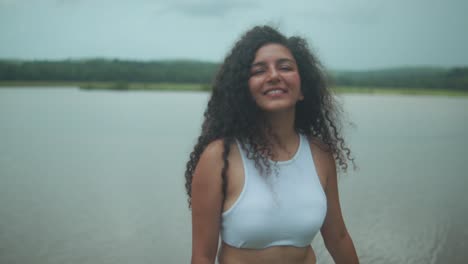 a young woman in a white outfit smiles and moves gracefully by the sea, with forested land in the background