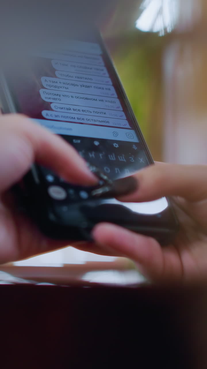 primer plano de un teléfono móvil negro que se utiliza para chatear en una mesa de madera, dedos escribiendo en el teclado, pantalla que muestra mensajes de texto, efectos de luz borrosos alrededor de la creación de un ambiente suave y cálido