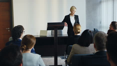 oradora de negocios caucásica en un podio en una conferencia e interactuando con personas en el salón durante su discurso