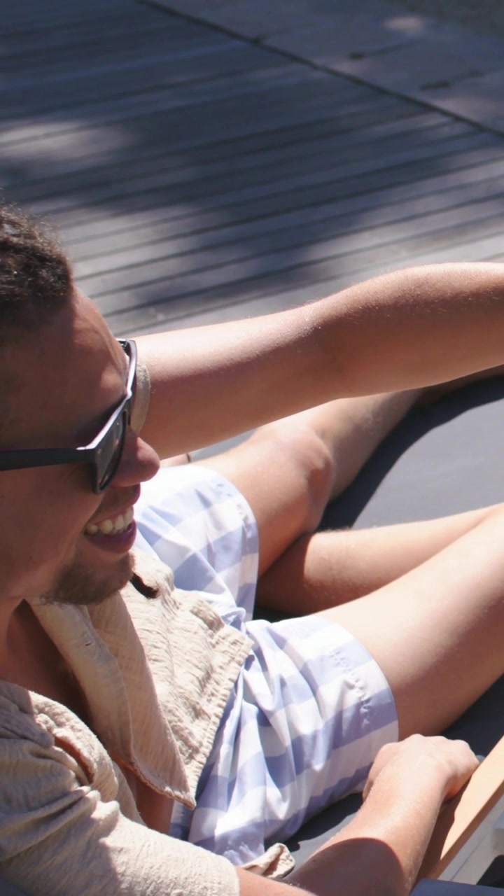 Vertical video: Glancing man in shades, open shirt, trunks reaching glass on deck, sipping to cool