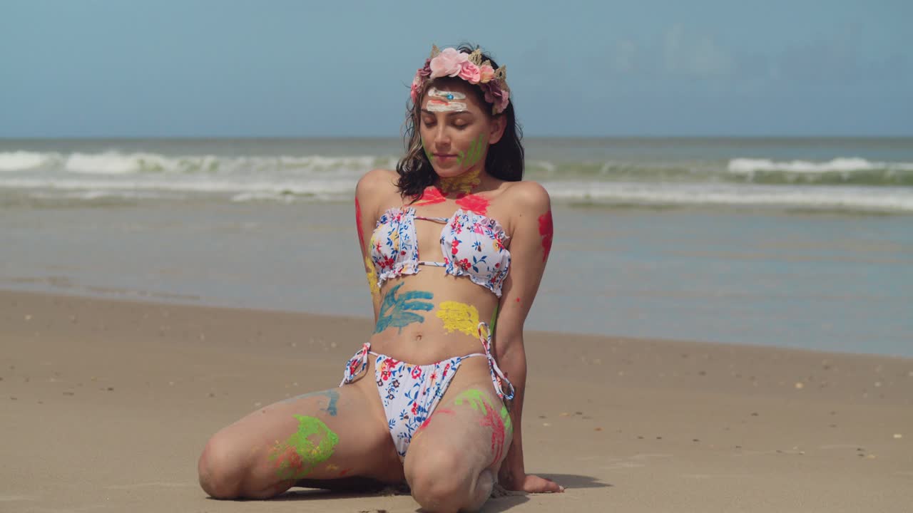 On a bright day in the Caribbean, a bikini-clad young girl with body paint kneel on the sandy shores