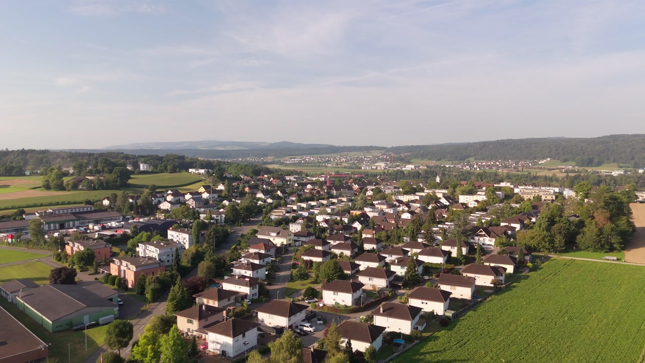 Forward drone reveals Stetten suburb bordered by fields and hills under clear summer skies.