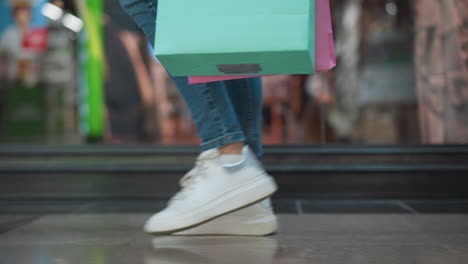 side leg view of a woman in jeans and canvas sneakers carrying a mint and pink shopping bag, walking in a well-lit shopping mall with her reflection visible on the glass