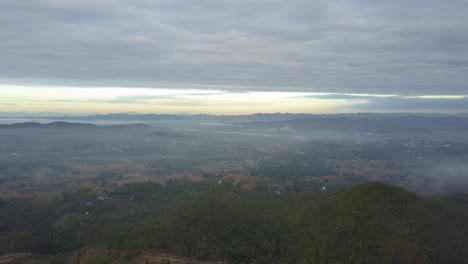 Vista-Del-Cielo-De-La-Ciudad-En-Tailandia-En-El-Fondo-Con-Un-Ambiente-Brumoso,-Montañas-Verdes-Y-Casas---Toma-Aérea