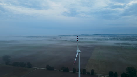 4k aerial: calm wind turbine and mystic fog hovering over farm fields in poland early in the morning before sunrise