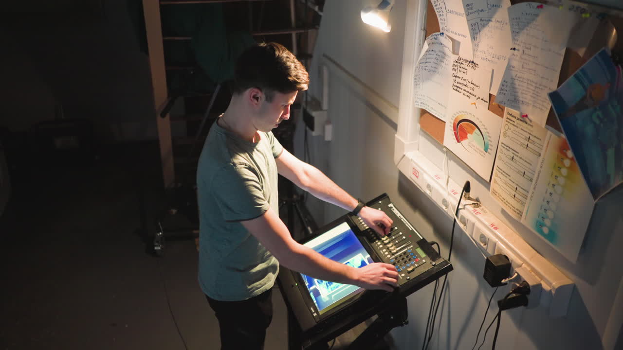 Young male technician stands under focused light in dark backstage environment, adjusting digital control panel with touchscreen interface. Wall filled with handwritten notes
