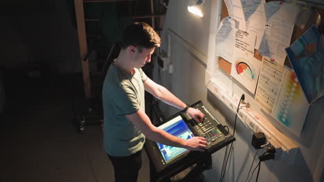 young male technician stands under focused light in dark backstage environment, adjusting digital control panel with touchscreen interface. wall filled with handwritten notes