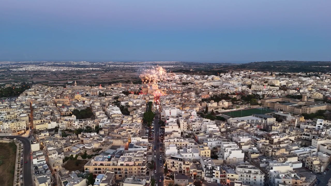 Rabat, Malta, Basilica di San Paolo e piazza pubblica decorata durante una festa di paese al crepuscolo, con fuochi d'artificio, luci della città e architettura mediterranea storica in vista aerea