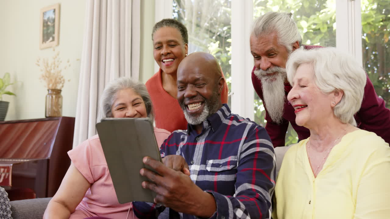 Amigos idosos diversos e felizes, homens e mulheres, usando tablet juntos em uma sala de estar ensolarada, em câmera lenta.