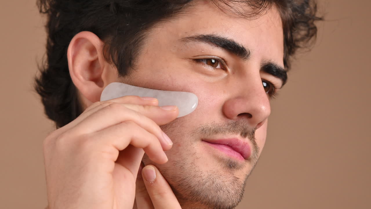 A young caucasian man with stubble beard is doing a facial massage using a Gua Sha