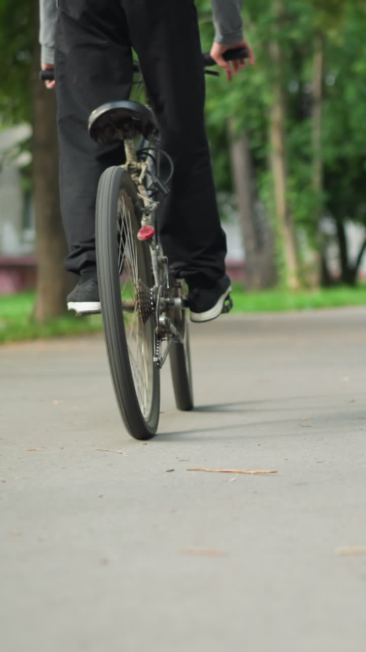 검은 스니커와 바지를 입은 사이클리스트의 뒷면, 초록색으로 둘러싸인 자전거 근처에 서 있는 다른 개인의 흐릿한 배경과 함께 포장 된 길을 따라 꾸준히 자전거를 페달