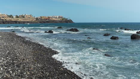 Pequeñas-Olas-Del-Océano-Rompen-Suavemente-En-Una-Playa-Rocosa-De-Guijarros-Con-Hoteles-Junto-A-La-Playa-En-El-Fondo