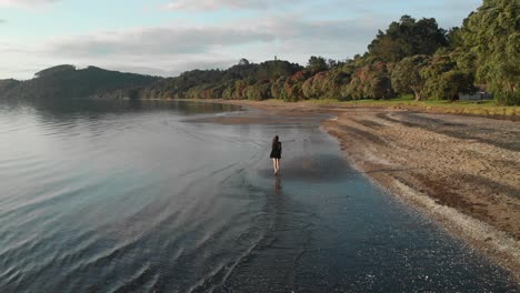 Aerial-drone-view-of-girl-running-Cornwallis-beach-in-Auckland,-New-Zealand,-following-shot