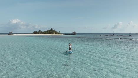 fit woman on standup paddleboard rowing towards tropical island in maldives