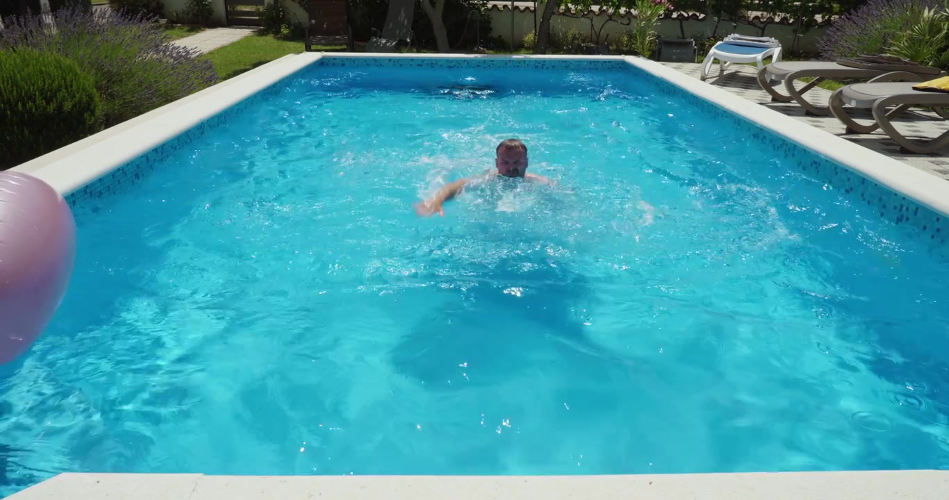 Mand svømmer på tværs af en pool, mens han øver sin svømmeteknik i slowmotion. Koncept for fitness, udholdenhed, vandtræning og sund livsstil på en solrig sommerdag.
