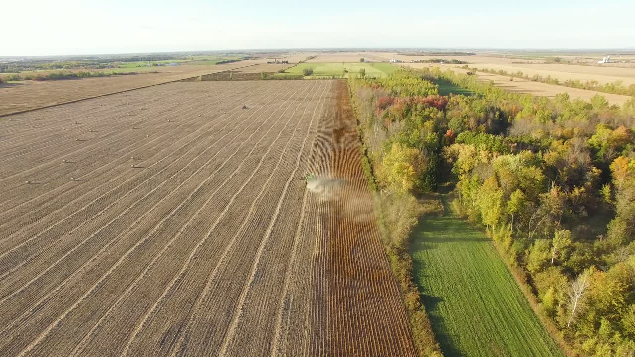 High angle, aerial view of dust rising behind farm equipment working in a brown open field. Wide, flat terrain with farms and trees on the horizon in Autumn.