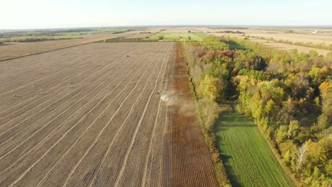 high angle, aerial view of dust rising behind farm equipment working in a brown open field. wide, flat terrain with farms and trees on the horizon in autumn.