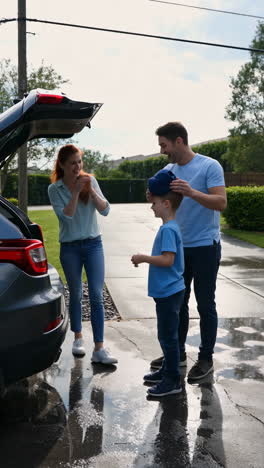 a family stands next to an open car trunk in a driveway. the parents smile at their young son, who adjusts his baseball cap