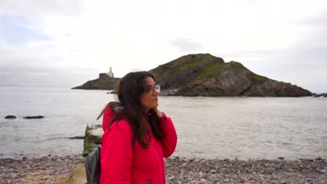 Mujer-Con-Chaqueta-Roja-Sonríe-Por-La-Bahía-De-Pulsera-Con-Faro-E-Islas-De-Murmullos-En-El-Fondo,-Gales